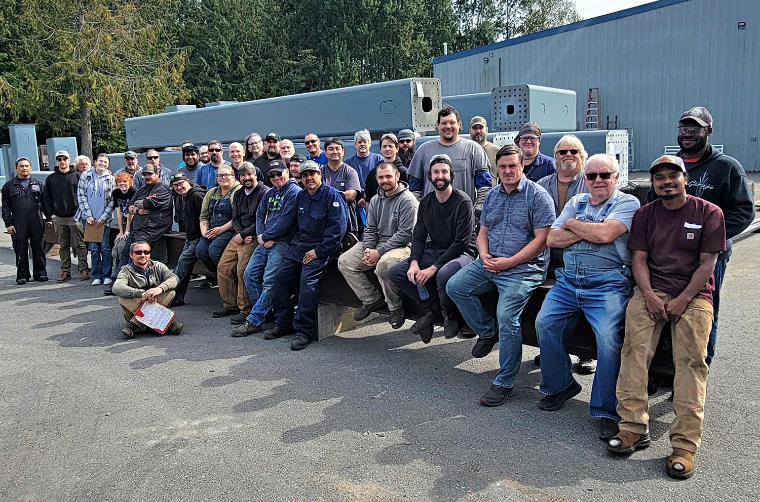 A large group of EDCO, INC. team members posing outdoors in front of industrial components and a manufacturing facility, with trees in the background.